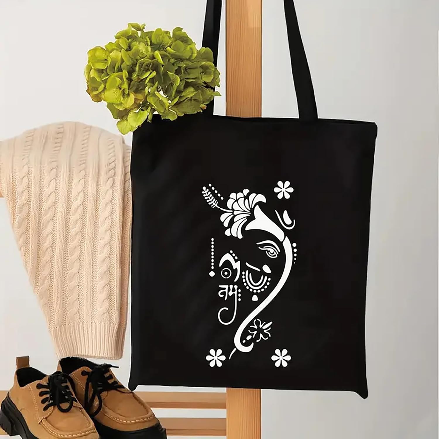 Black tote bag with white floral design, beige sweater, and brown shoes on a light gray background