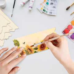 Person painting sunflowers on a card with art supplies on a table.