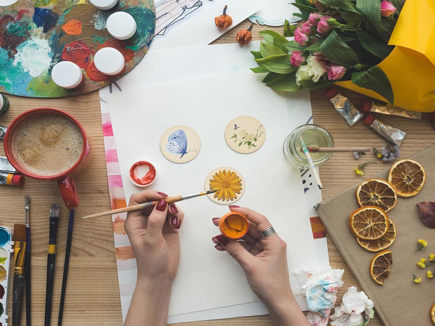 Person painting a small round object with a brush on a table with art supplies and flowers.