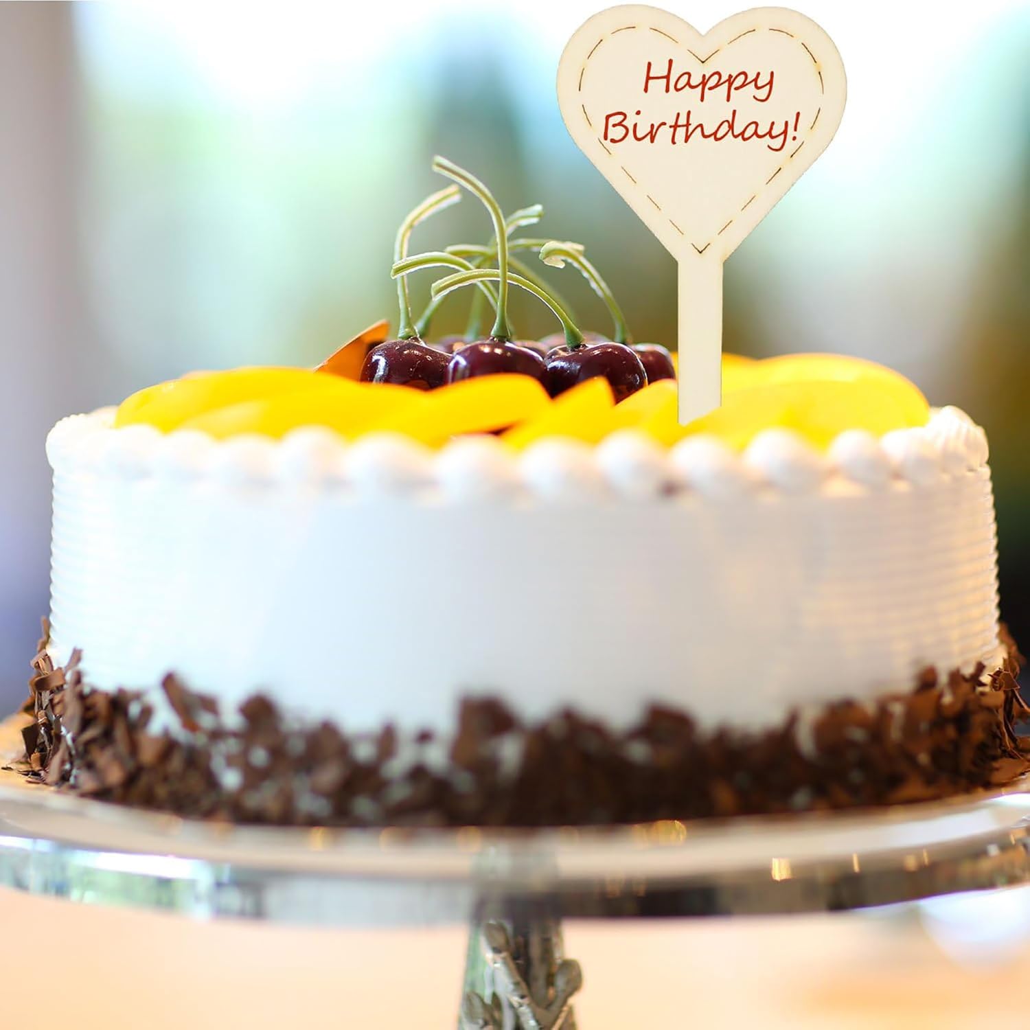 White birthday cake with fruit toppings and a 'Happy Birthday!' sign on a blurred background