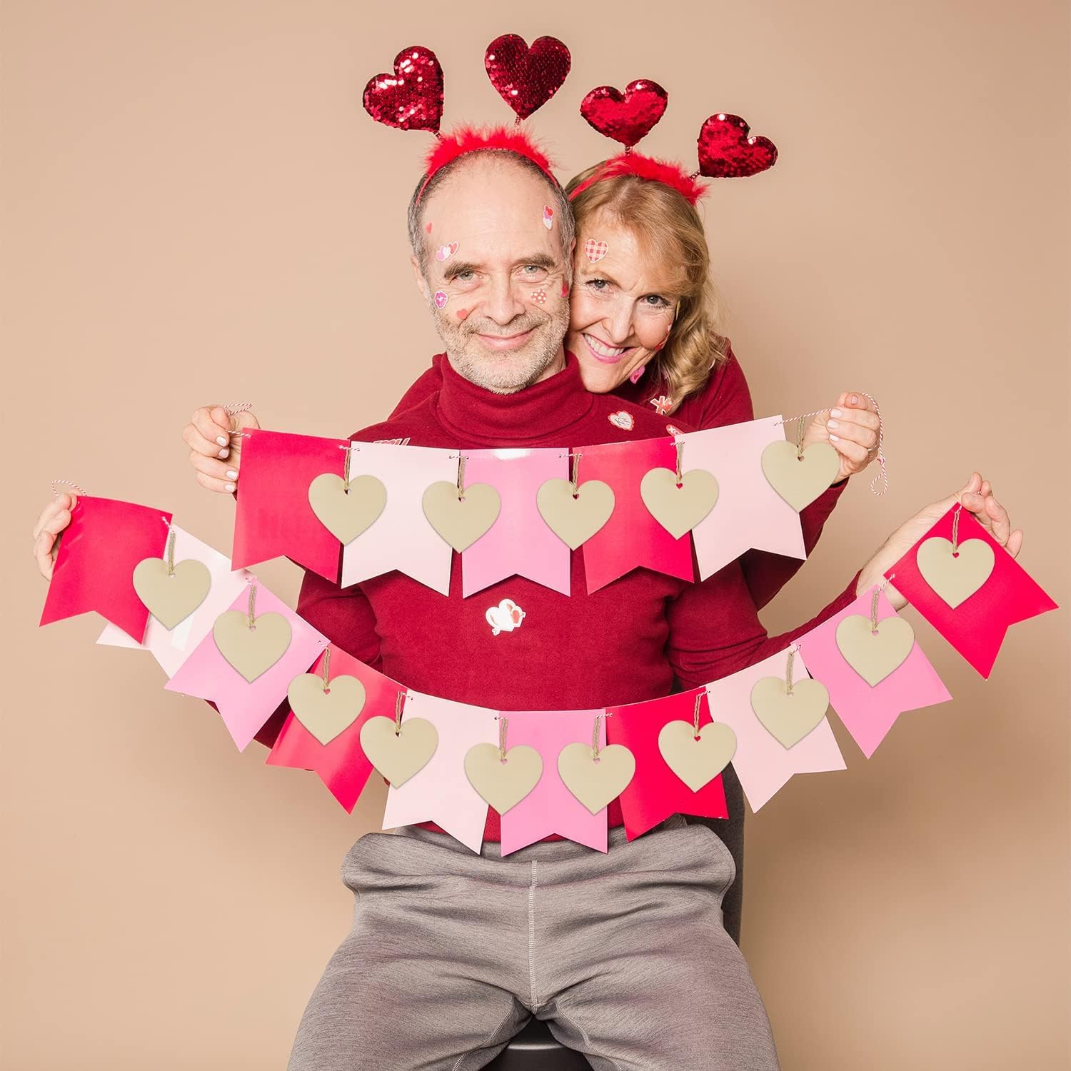 Two people wearing heart-themed headbands and holding a heart-shaped banner against a beige background