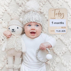 Baby holding a teddy bear and a teething ring with a 'Today I am 5 months old' sign on a textured white background.