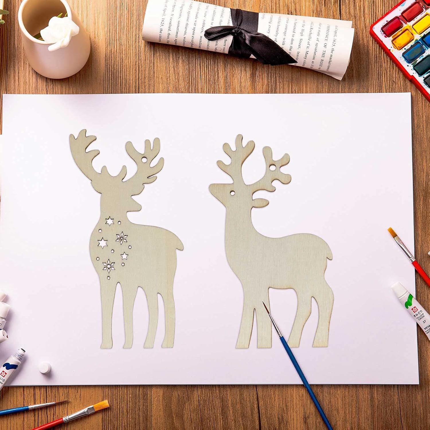 Two paper reindeer cutouts on a white sheet of paper with art supplies around.