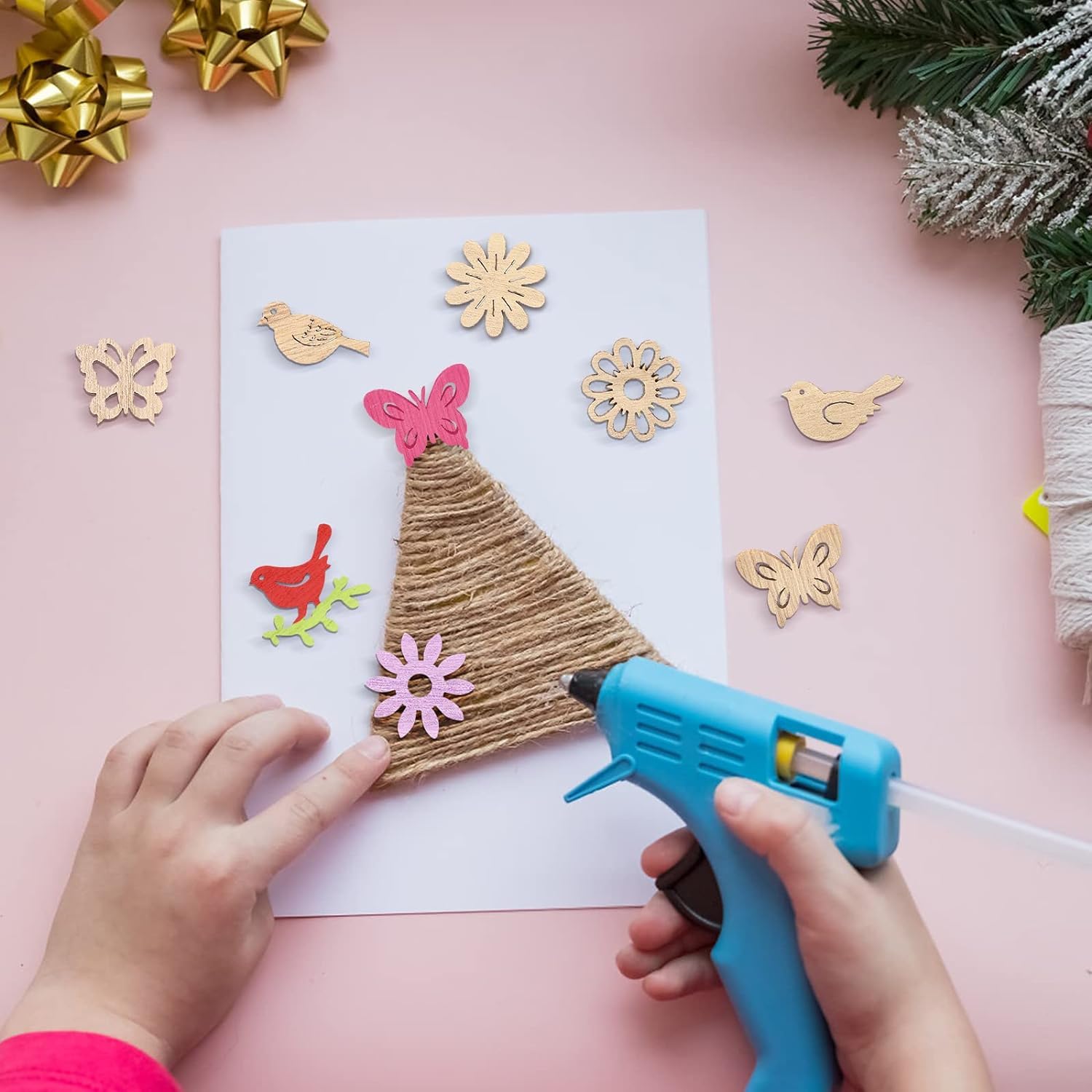 Child's hands using a hot glue gun to decorate a card with paper flowers and birds on a pink background.
