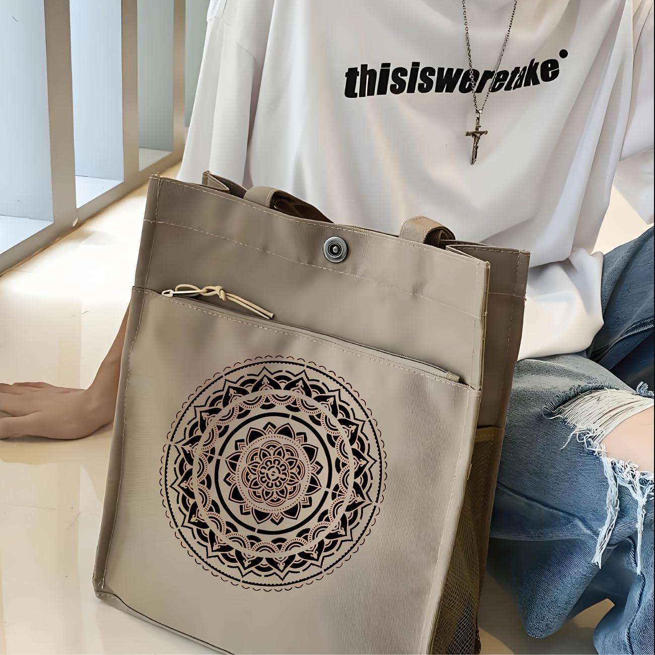 Beige tote bag with mandala design held by a person wearing a white shirt with text.