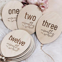Wooden milestone discs labeled 'one month', 'two months', 'three months', and 'twelve months' on a soft surface with flowers.