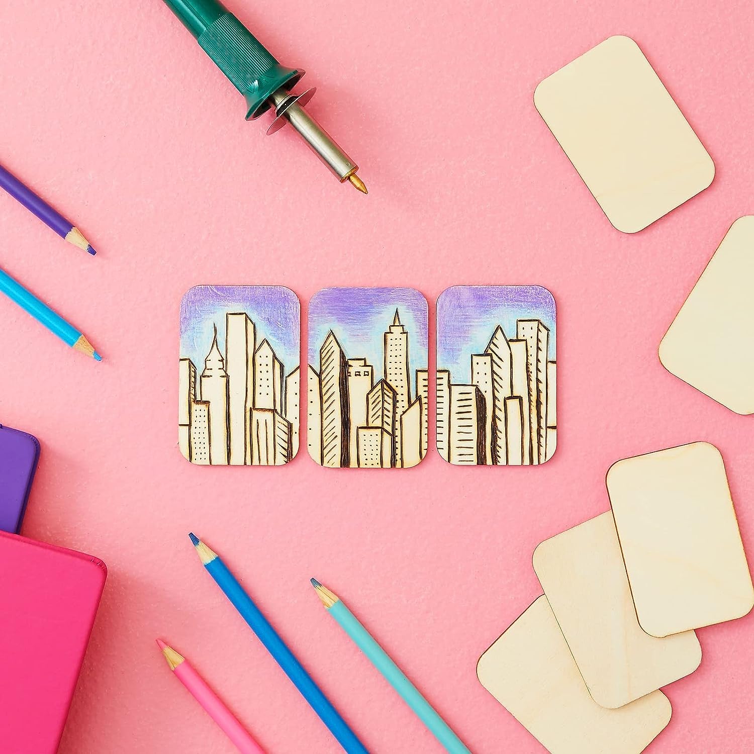 Three wooden coasters with cityscape design on a pink background with art supplies.
