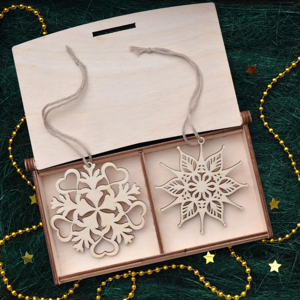 Two wooden snowflake ornaments in an open box on a green textured background with gold decorative elements.