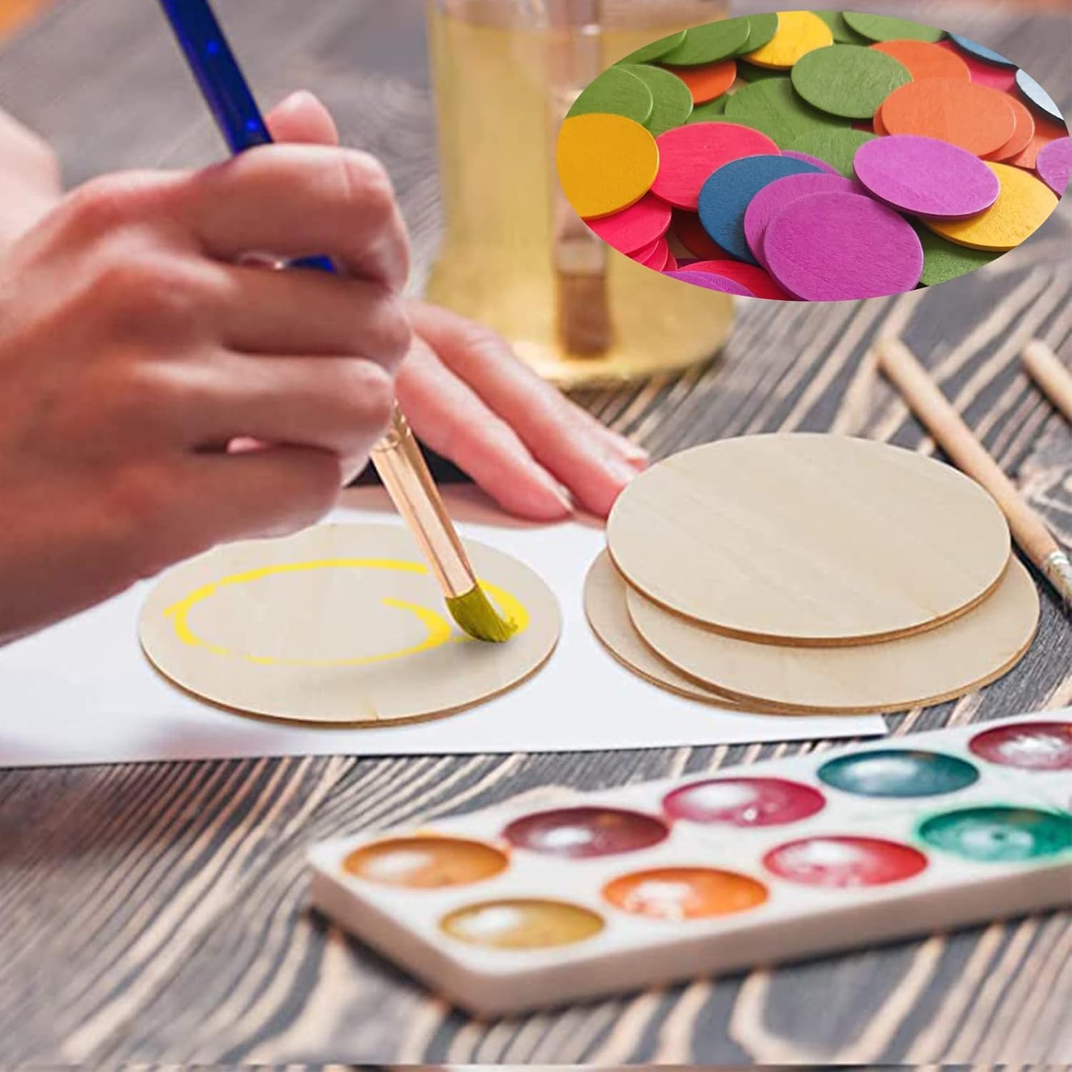 Person painting wooden circles with watercolors on a wooden table, surrounded by paint supplies.