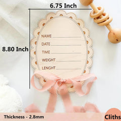 Wooden baby milestone card with measurements and text space, accompanied by a pink ribbon and wooden rattle.