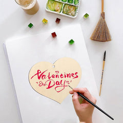 Heart-shaped card with 'Valentine's Day' writing being painted on a white surface with art supplies.