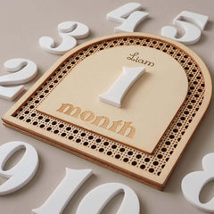 Wooden plaque with 'Liam' and '1 month' engraved, surrounded by white numbers on a beige background