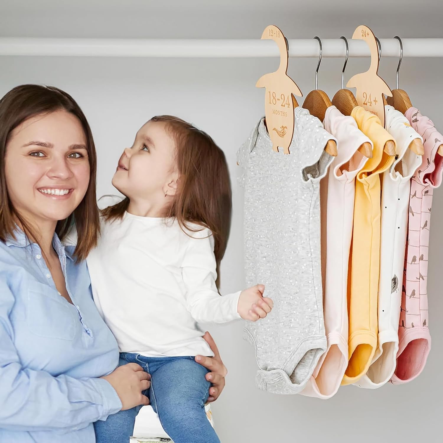 Woman holding a child next to a rack of colorful baby clothes on wooden hangers.