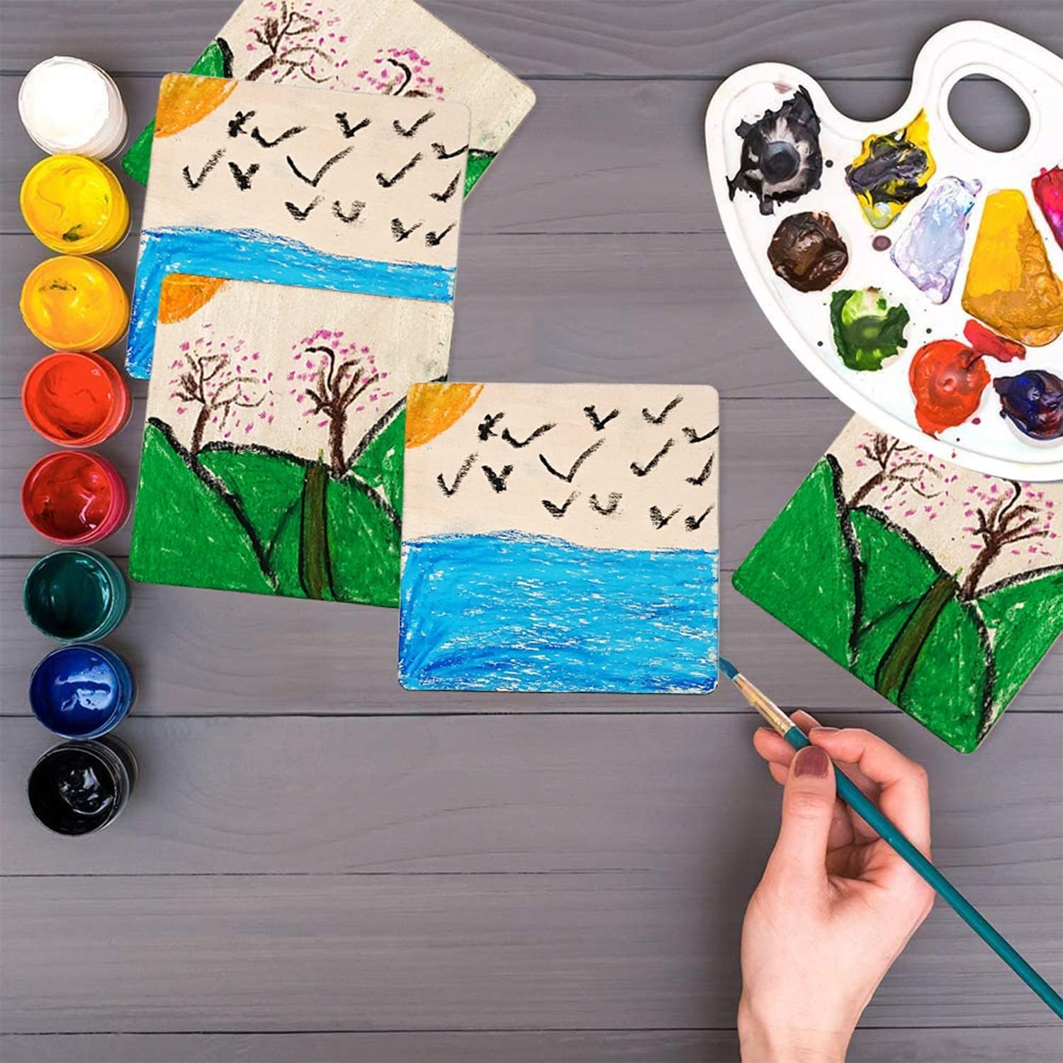Children's artwork with trees, birds, and water on a wooden surface, accompanied by paint supplies.