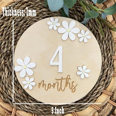 Wooden disc with '4 months' and white flowers on a woven surface
