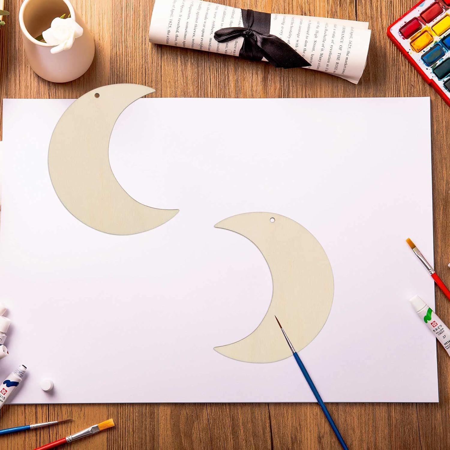 Two crescent moon shapes on a piece of paper with painting supplies on a wooden table.