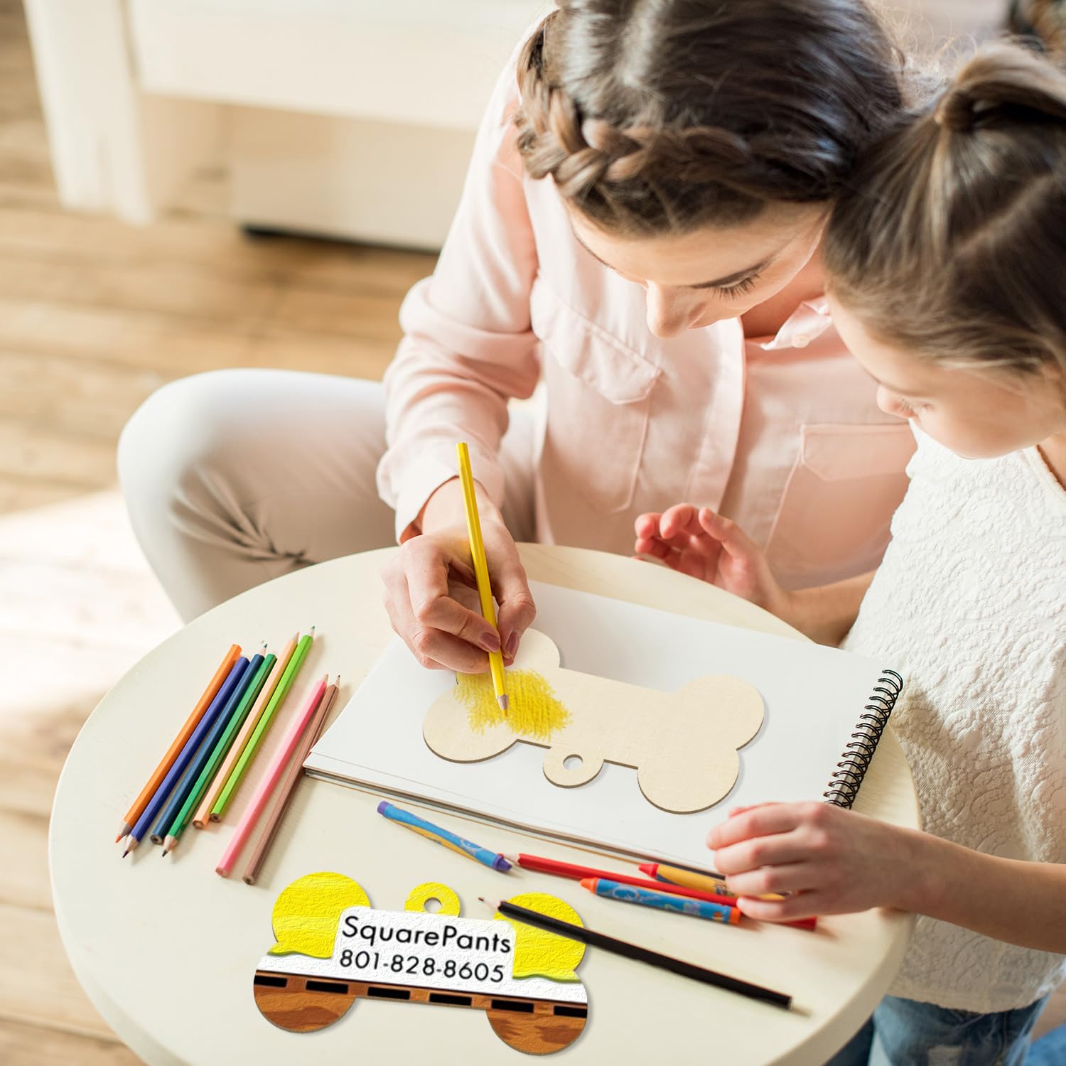 Two children sitting at a table with art supplies, including colored pencils and a wooden craft, with a SquarePants logo in the corner.
