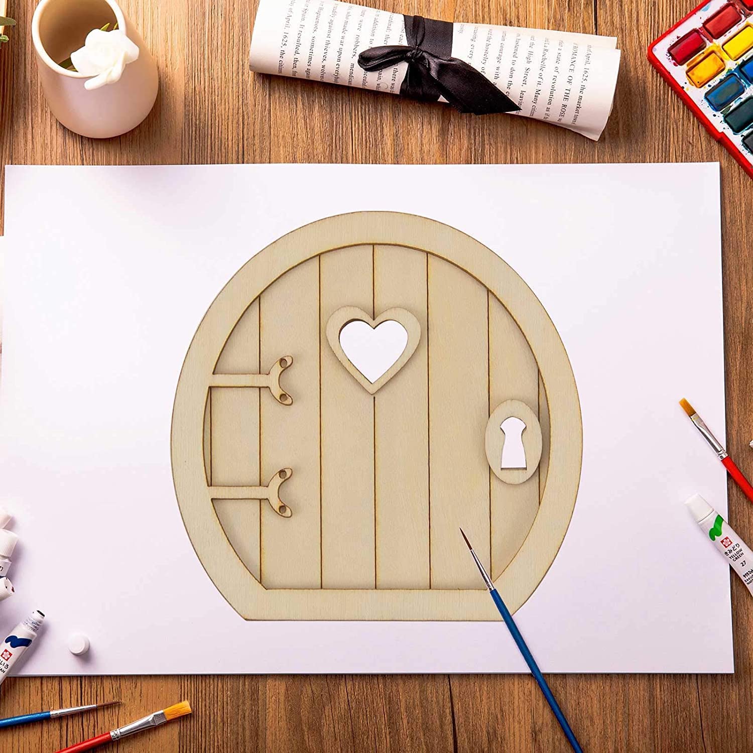 Wooden fairy door with heart-shaped window on a white sheet of paper, surrounded by art supplies on a wooden table.