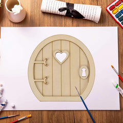 Wooden fairy door with heart-shaped window on a white sheet of paper, surrounded by art supplies on a wooden table.