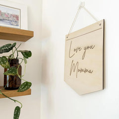 Wooden sign with 'Love you Momma' text hanging on a wall next to a plant and shelf.