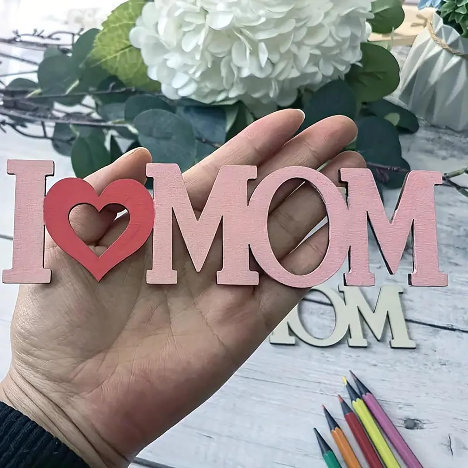 Hand holding wooden letters spelling 'I ❤️ MOM' with flowers and stationery in the background