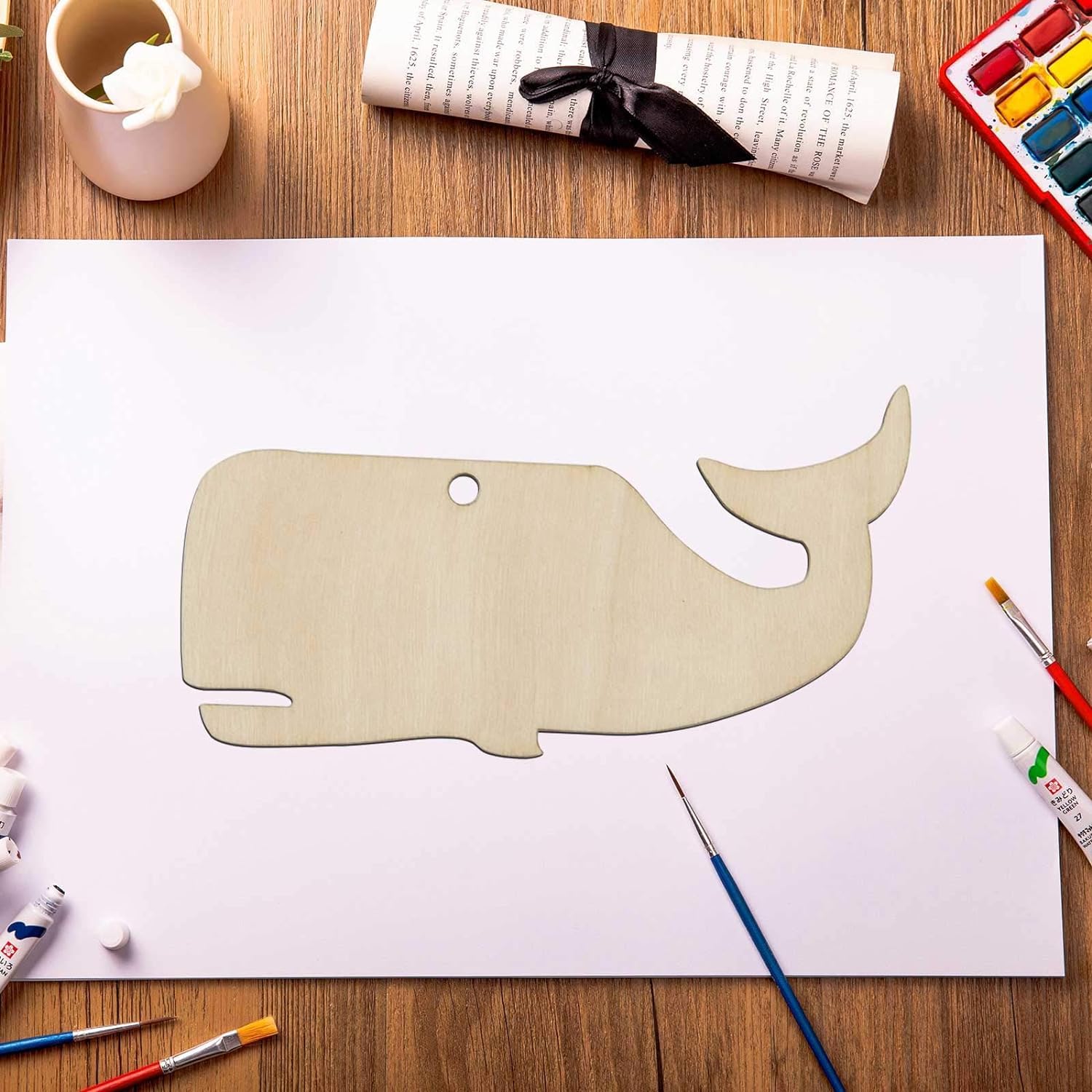 Wooden whale cutout on a white sheet of paper with art supplies on a wooden table.