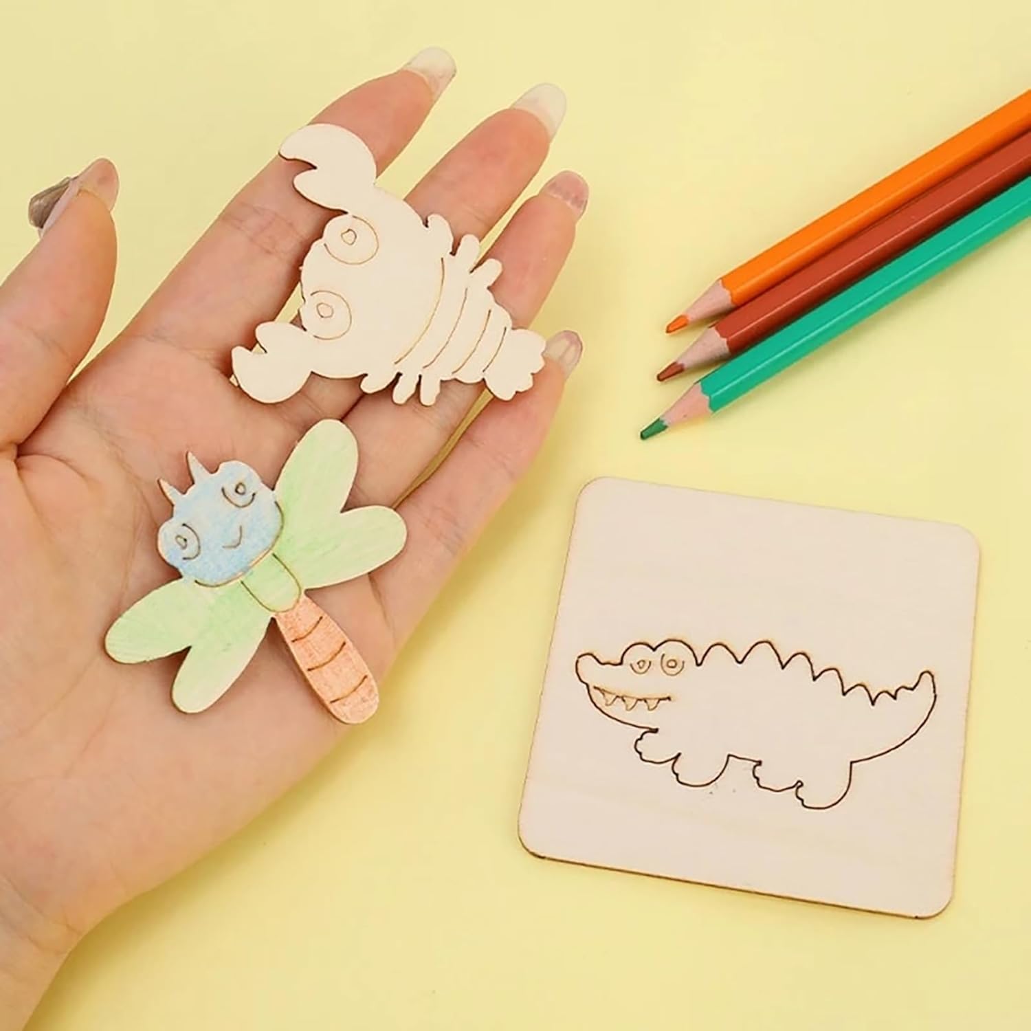 Wooden cutouts of a dragonfly and a crab held by a hand on a yellow background with coloring pencils.