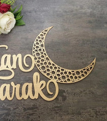 Decorative wooden crescent moon with 'Eid Mubarak' text on a textured surface with flowers.