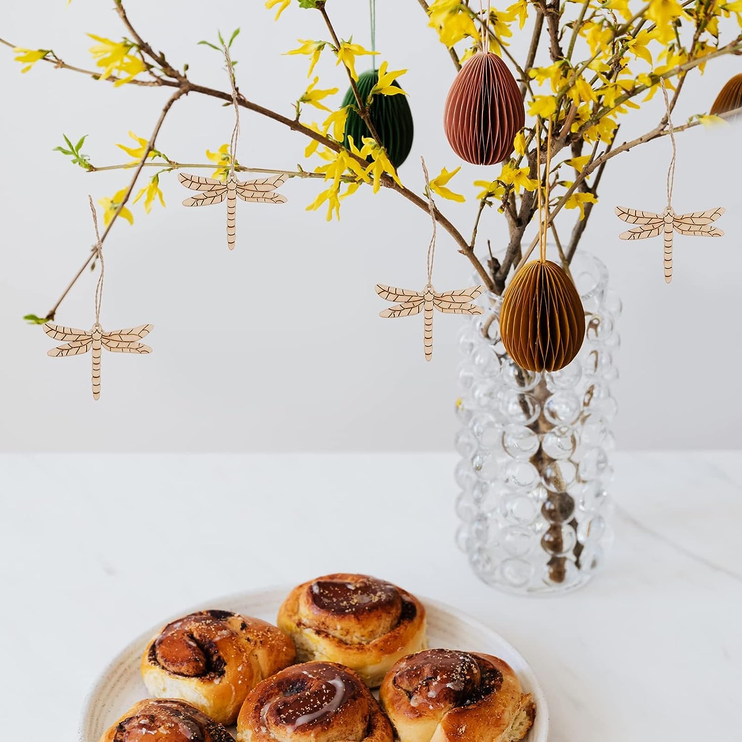 Decorative Easter eggs and dragonflies hanging from a branch with yellow flowers, next to a plate of cinnamon rolls.