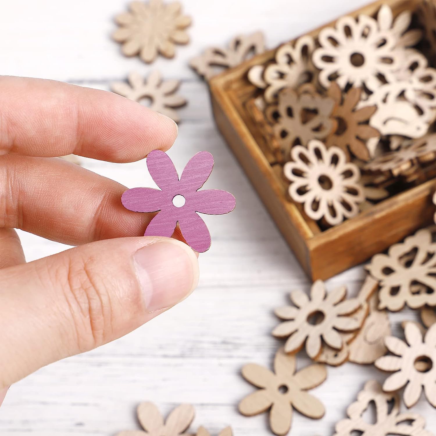 Hand holding a purple flower-shaped wooden cutout with more similar cutouts on a white surface.
