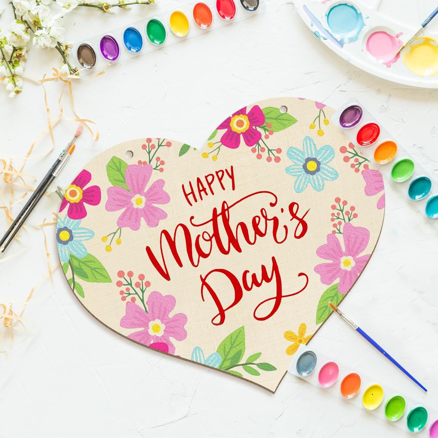 Heart-shaped sign with 'Happy Mother's Day' surrounded by flowers, paintbrushes, and paint on a white background