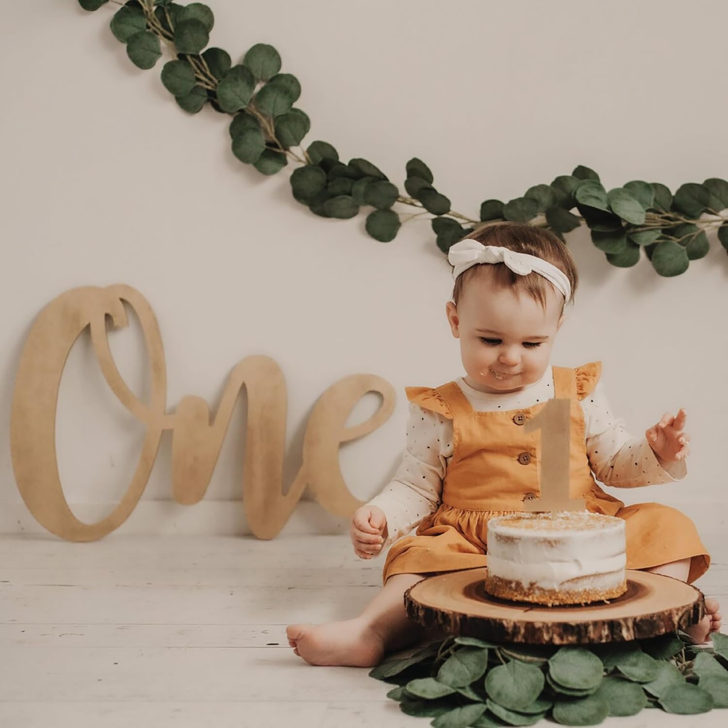 Baby in an orange dress sitting next to a cake with 'One' wooden letters on a white background.