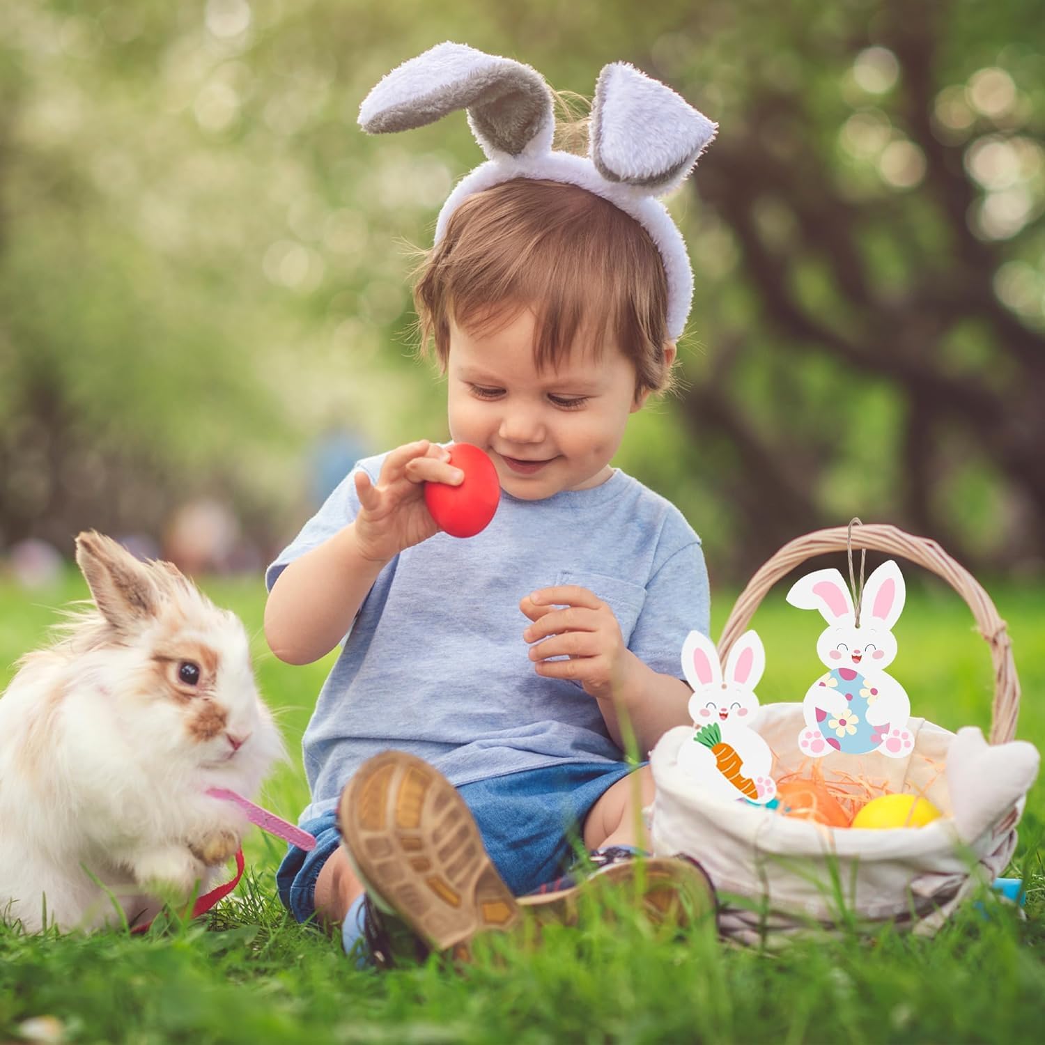 Child wearing bunny ears playing with a red egg in a grassy field with a rabbit and Easter basket.
