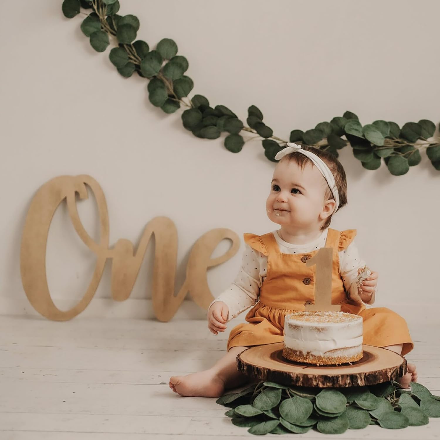 Baby in a yellow dress with a small cake and 'One' sign, surrounded by greenery.