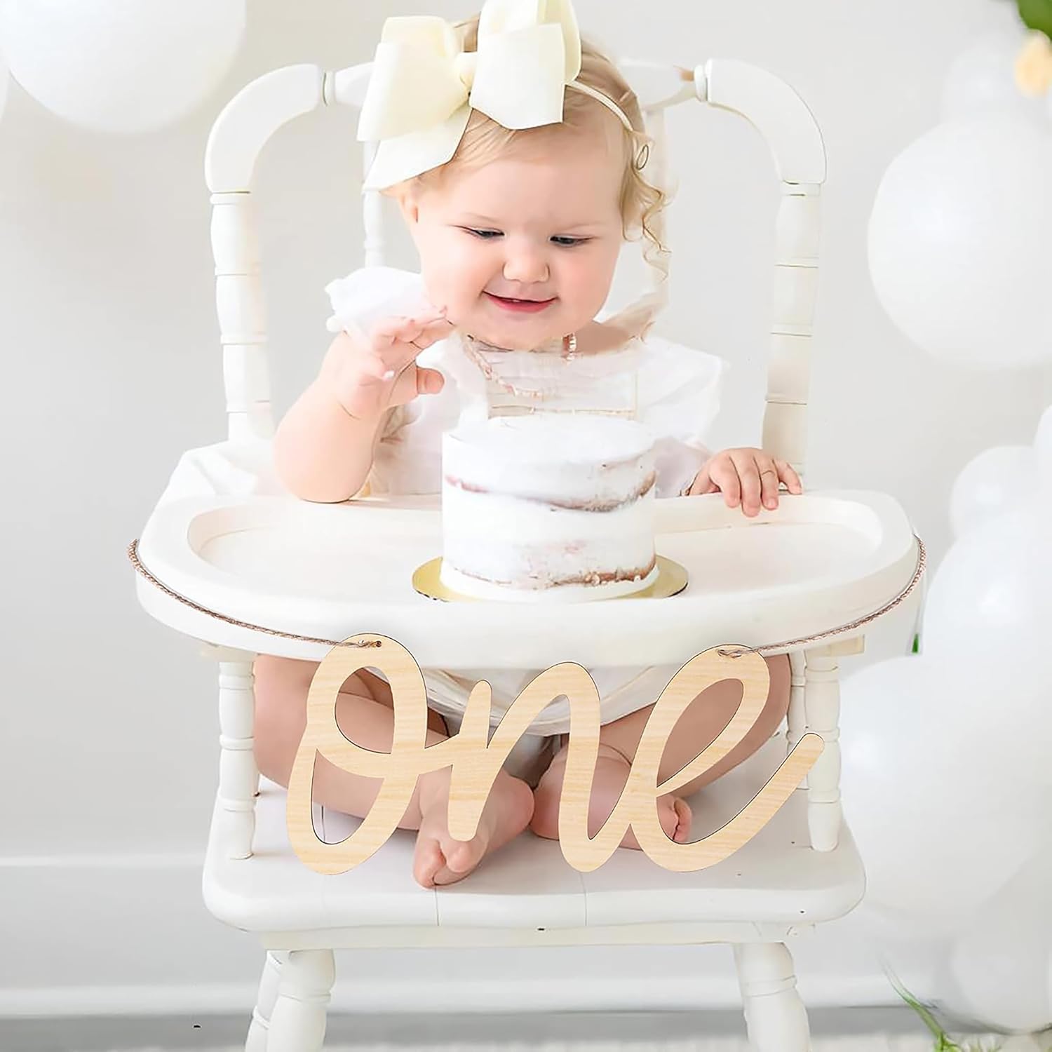 Baby in a high chair with a small cake and 'ONE' sign, wearing a white outfit and headband.