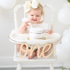 Baby in a high chair with a small cake and 'ONE' sign, wearing a white outfit and headband.