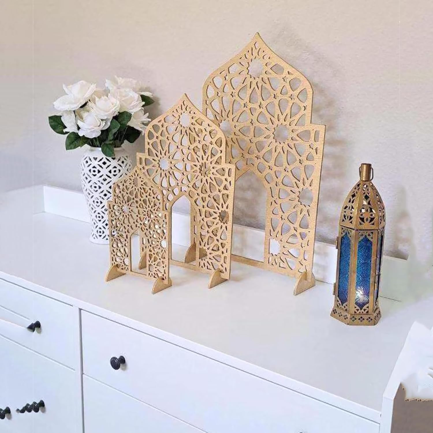 Decorative wooden arches and a lantern on a white surface with a vase of flowers in the background.