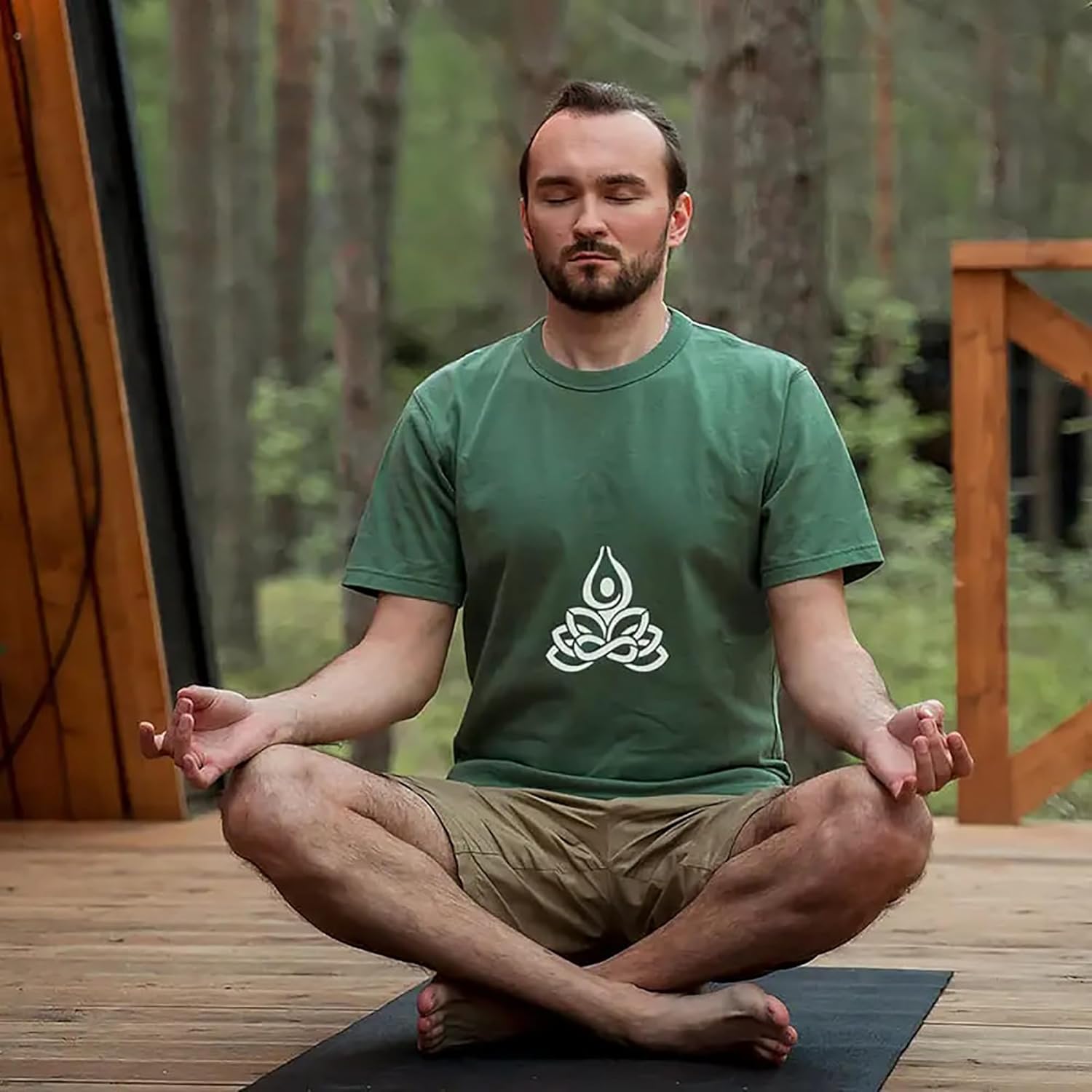 Man in a green t-shirt with a logo sitting in meditation on a wooden deck.