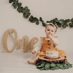 Baby in an orange dress sitting next to a small cake with a 'one' candle, surrounded by greenery.