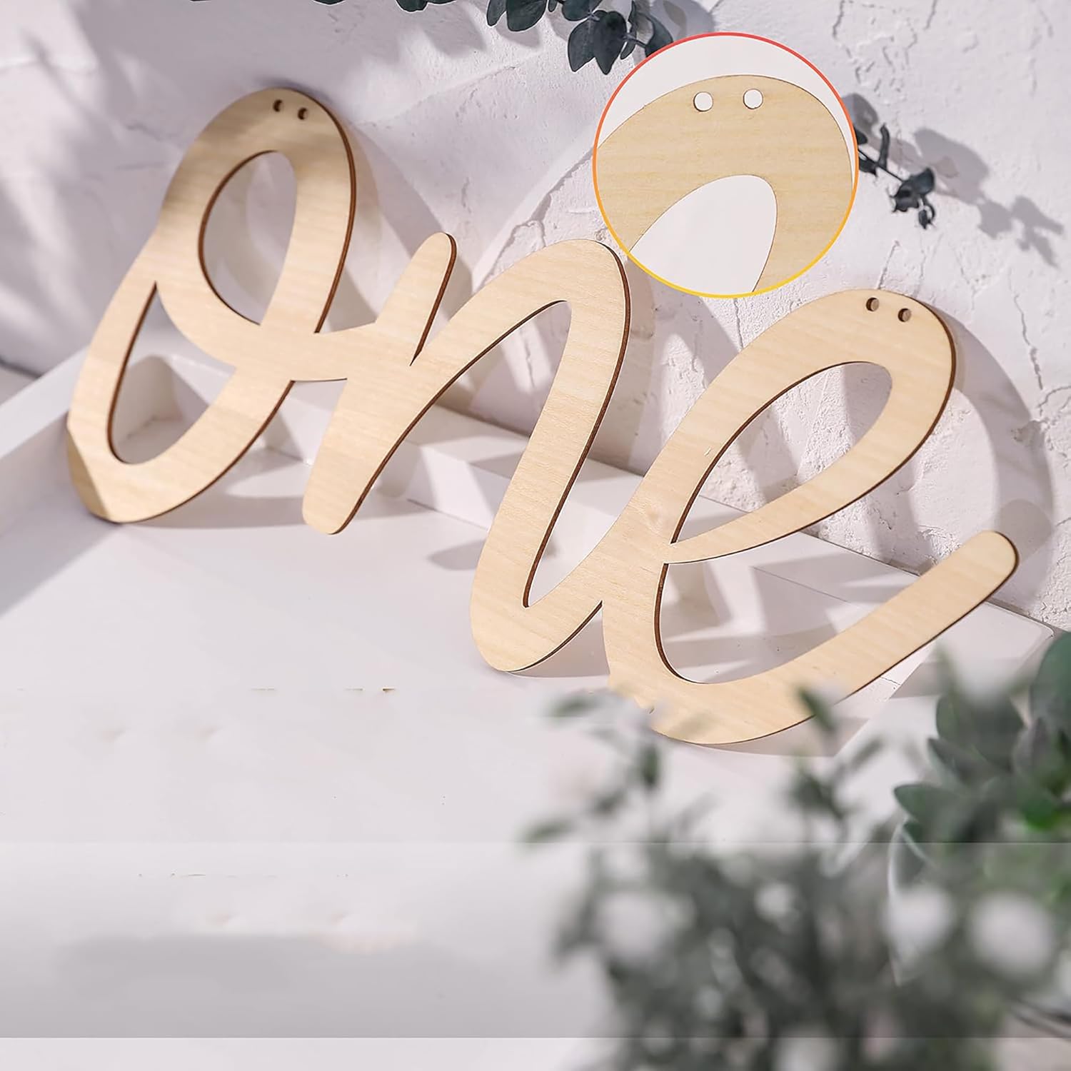 Wooden letter 'One' decoration on a white surface with a blurred green plant in the background.