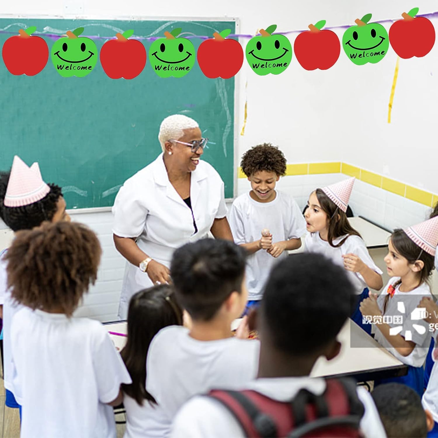 Teacher and students in a classroom with a 'Welcome' banner and apple decorations.