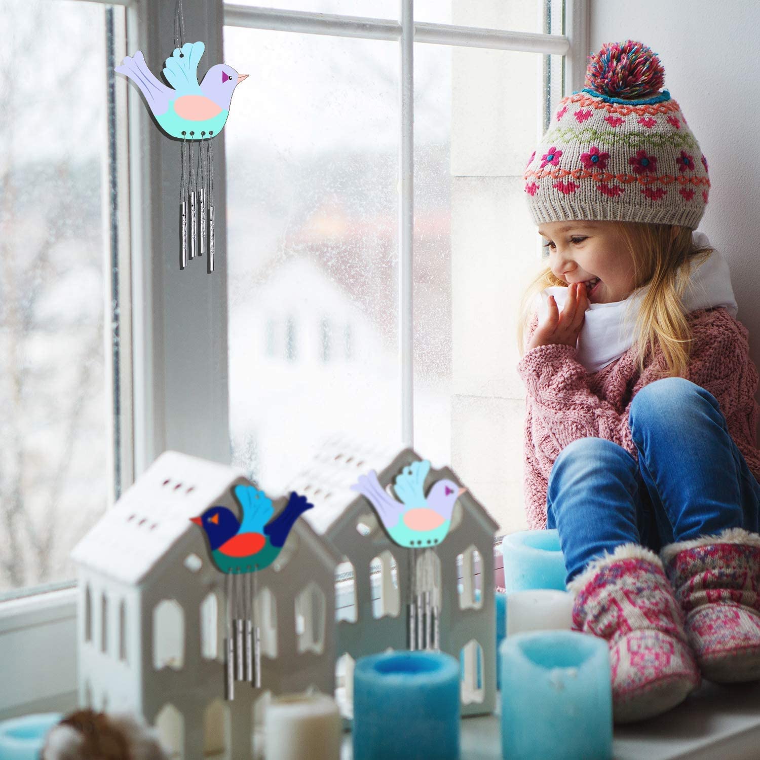 Child in winter clothing sitting by a window with decorative birdhouses and candles.