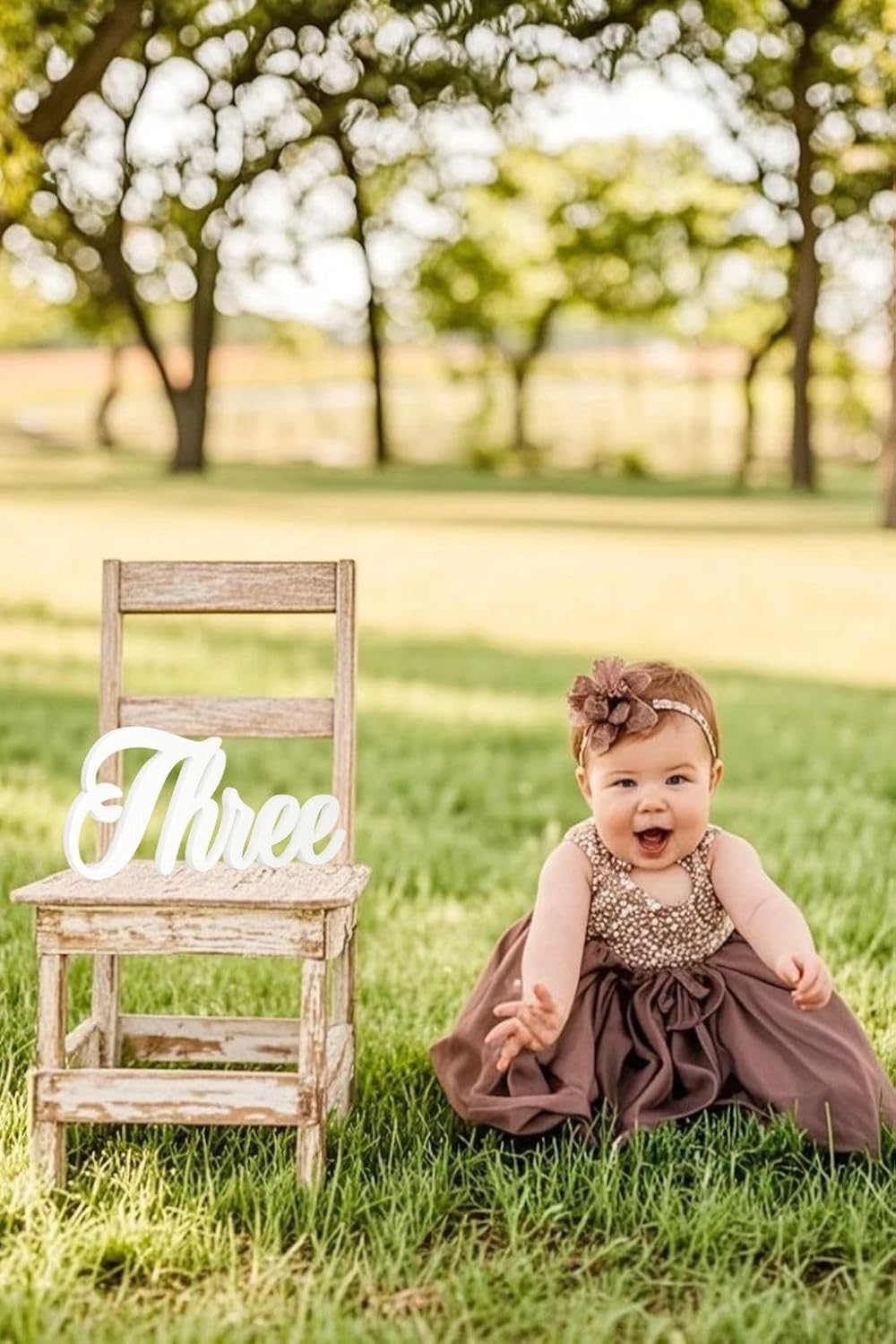 Baby in a dress sitting on grass next to a chair with 'Three' sign