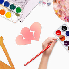 Hand painting a heart with watercolors on a white surface with paint supplies.