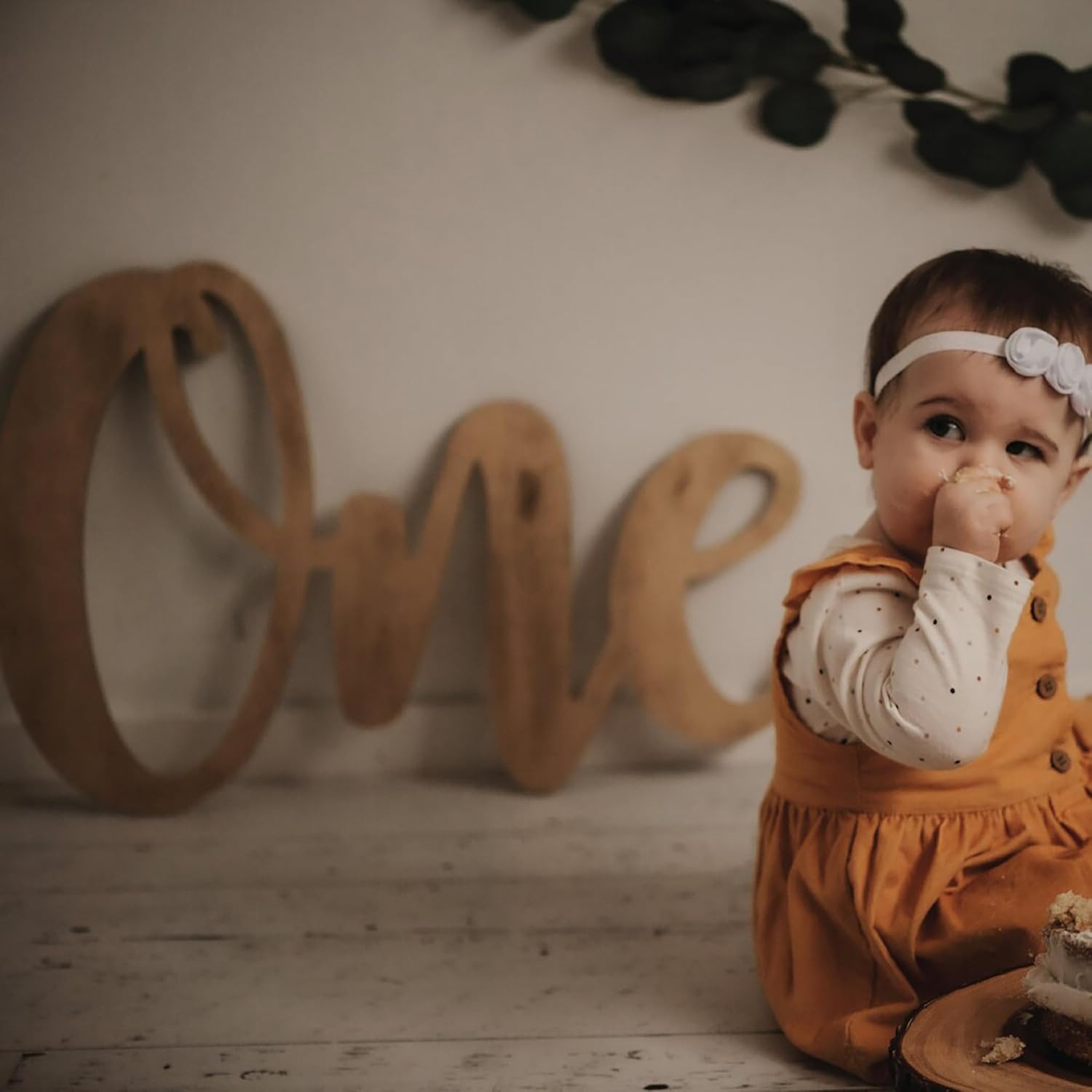 Baby in an orange outfit with a white headband, sitting in front of a wooden 'One' sign.