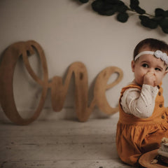Baby in an orange outfit with a white headband, sitting in front of a wooden 'One' sign.