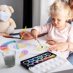 Child painting with watercolors at a table, assisted by an adult.