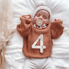 Newborn baby wearing a knitted brown sweater with '4 months' on it, lying on a soft white surface.