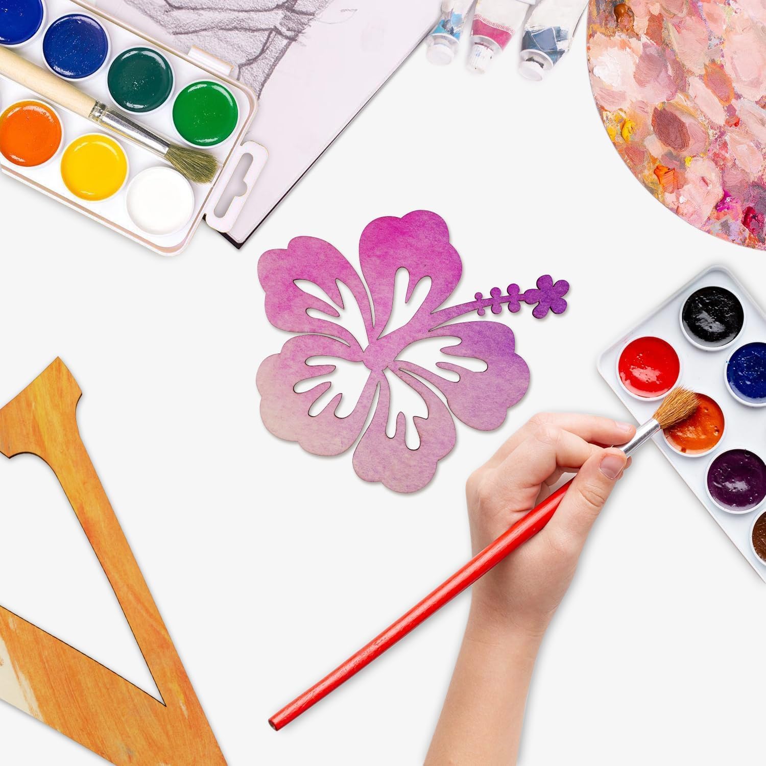 Artistic activity with watercolor paints, brushes, and a hand painting a flower design on a white surface.