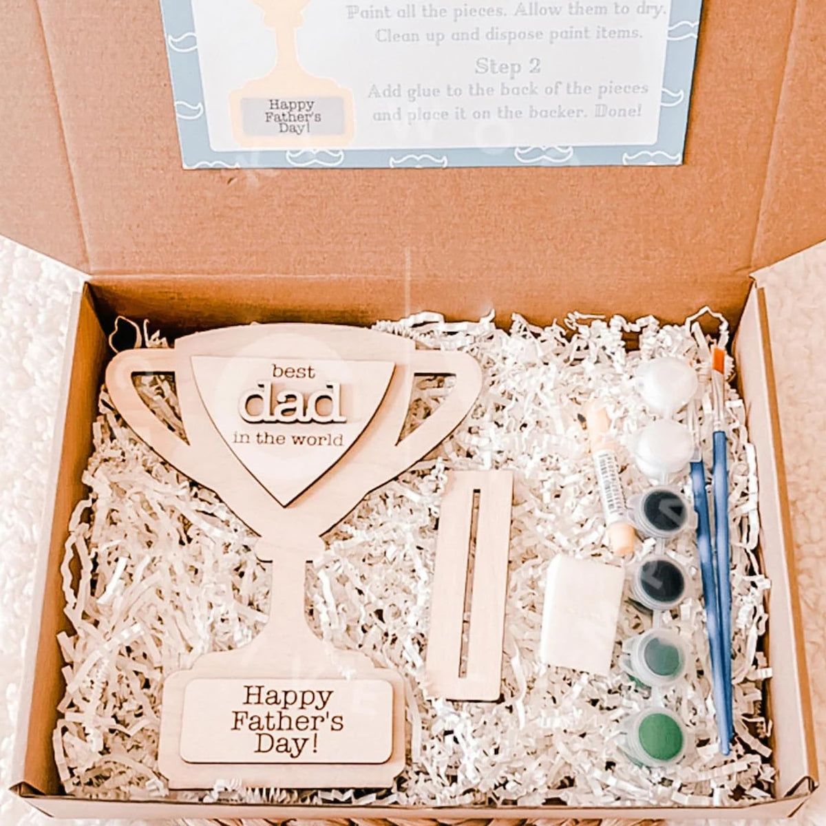 Wooden 'best dad in the world' plaque with paint supplies in a box, perfect for Father's Day.